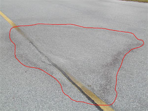 Photo showing a medium-severity depression on the pavement.  The area of the           depression is noticeable due to water staining on the pavement.  A red line has also been drawn on the photo to outline the area with the depression.