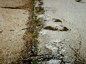 Close-up photo showing a longitudinal joint between a PCC and an asphalt pavement.            The asphalt surface in the photo has a considerable amount of distress that has occurred from shoving.